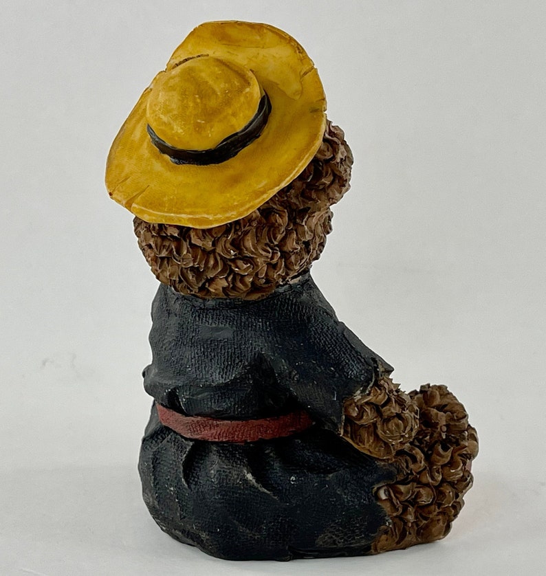 May include: A brown teddy bear figurine wearing a yellow straw hat with a black band and a blue jacket with a red belt. The bear is sitting with its back to the camera.