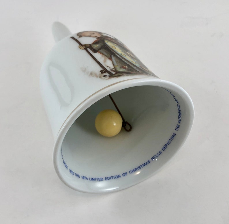 May include: White porcelain bell with a blue and gold trim. The bell features a painting of a girl in a long dress holding a stick. The inscription on the bell reads "1974 Limited Edition of Christmas Bells Depicting the Authentic Art".
