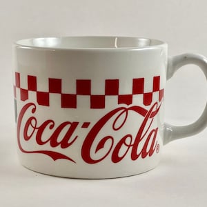 Op de afbeelding: Witte keramische Coca-Cola mok met een rood geblokt patroon en het Coca-Cola logo in rood. De mok heeft een gebogen handvat en een klassiek ontwerp. Een geweldige aanvulling op elke keuken.