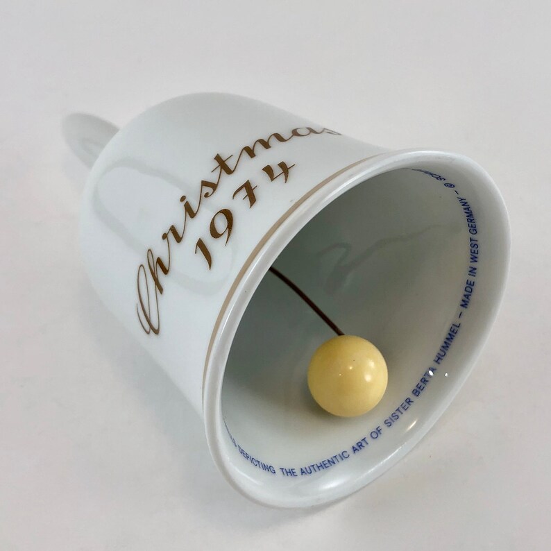 May include: White porcelain Christmas bell with gold lettering that reads "Christmas 1974". The bell has a small, yellow, round clapper inside.
