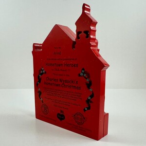 May include: Red wooden Christmas ornament shaped like a house with a white inscription that reads "Part No. A1518 In the spirit of appreciation of Hometown Heroes Third Edition in the Charles Wysocki's Hometown Christmas This ornament is produced in a limited, hand-numbered series. Each ornament is crafted of the finest cast resin, exacting the highest standards of quality. The officially licensed Charles Wysocki's Hometown Christmas 2021 Ornament Series"