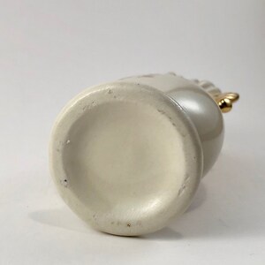 May include: White ceramic vase with a gold handle. The vase has a smooth, rounded bottom.