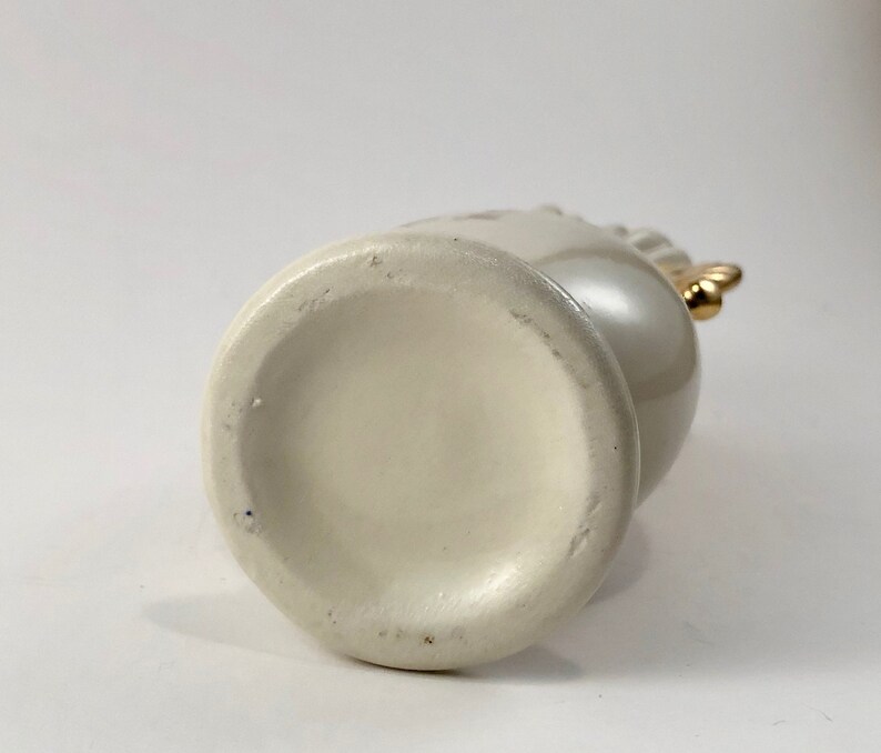 May include: White ceramic vase with a gold handle. The vase has a smooth, rounded bottom.