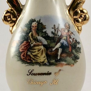 May include: A white ceramic vase with gold accents and a painted scene of two people in a garden. The vase has a gold inscription that reads "Souvenir of Chicago Ill."