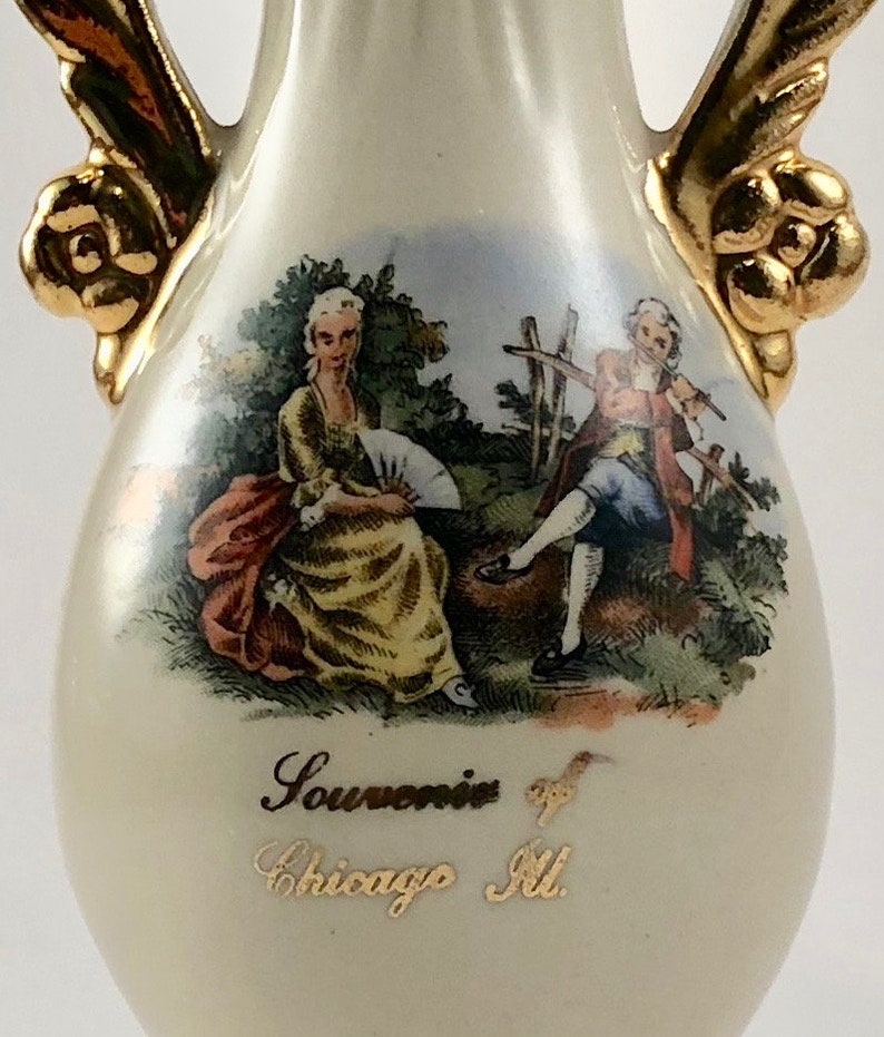 May include: A white ceramic vase with gold accents and a painted scene of two people in a garden. The vase has a gold inscription that reads "Souvenir of Chicago Ill."