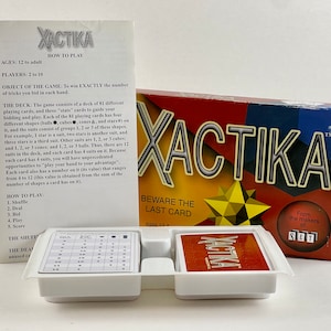 May include: XACTIKA card game box set with instructions and playing cards. The box is red, blue, and yellow with the game's name in large letters. The game includes cards and instructions.