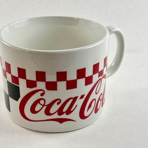 Op de afbeelding: Witte keramische mok met een rood-zwart geblokt patroon en het Coca-Cola-logo in rood. De mok heeft een gebogen handvat en een eenvoudig ontwerp, perfect voor dranken.
