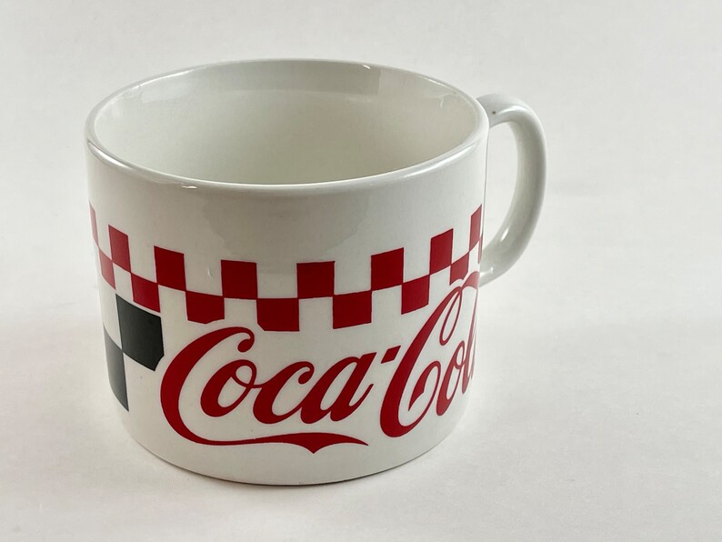 Op de afbeelding: Witte keramische mok met een rood-zwart geblokt patroon en het Coca-Cola-logo in rood. De mok heeft een gebogen handvat en een eenvoudig ontwerp, perfect voor dranken.