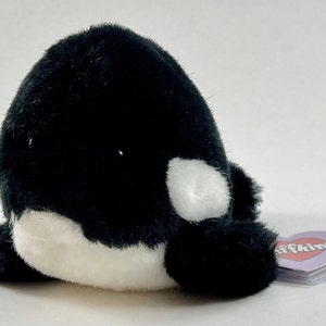 May include: Black and white plush orca stuffed animal with a white patch on its side. The orca is lying on its belly with its flippers outstretched. The orca has a pink heart-shaped tag that says "Soffkins" on it.