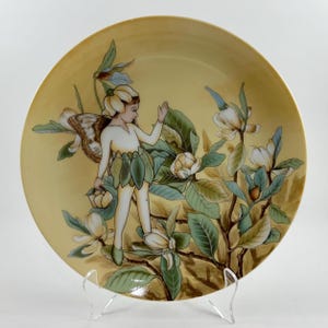 May include: A decorative plate with a fairy design. The plate features a fairy with a flower headdress and leaf skirt, standing among magnolia blossoms. The color palette includes yellow, green, brown, and white, creating a whimsical and artistic aesthetic.
