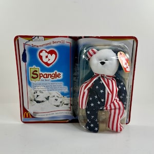 May include: A white Ty Beanie Baby named Spangle the Bear, wearing a red, white, and blue patriotic outfit. The bear is packaged in a clear plastic case with a red, white, and blue Ty Beanie Baby tag. The packaging features a red background with the words "International Bears II" and a picture of Mount Rushmore.