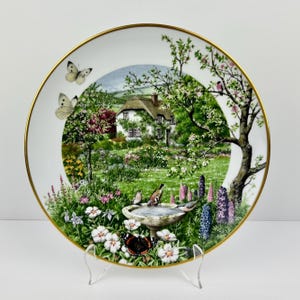 May include: Decorative plate featuring a detailed landscape scene with a cottage, garden, butterflies, and birds. The plate has a gold rim and is supported by a clear stand. The artwork showcases a vibrant garden setting.