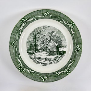 May include: A white ceramic plate with a green border and a black and white illustration of a farmhouse in a winter scene. The text "The Old Homestead in Winter" is printed on the plate.