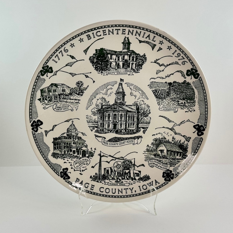 May include: A white ceramic plate with a black and green design commemorating the bicentennial of Page County, Iowa. The plate features illustrations of local landmarks, including the Amity College, the Page County Courthouse, and the Howell Mill. The text "1776 * Bicentennial * 1976" and "Page County, Iowa" are printed on the plate.