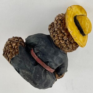 May include: A brown and black figurine of a bear wearing a black jacket with a red belt and a yellow hat.
