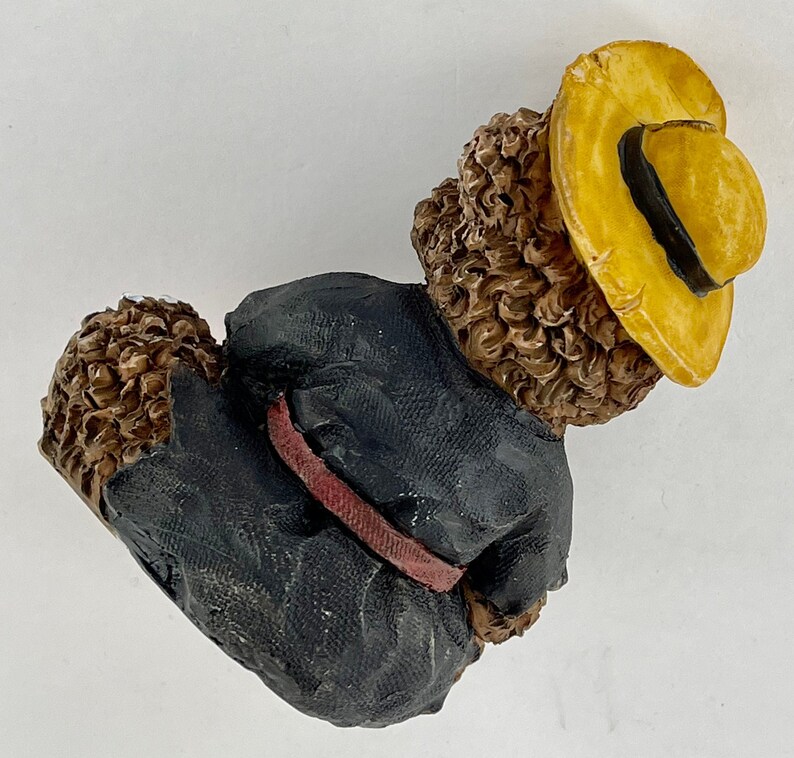 May include: A brown and black figurine of a bear wearing a black jacket with a red belt and a yellow hat.