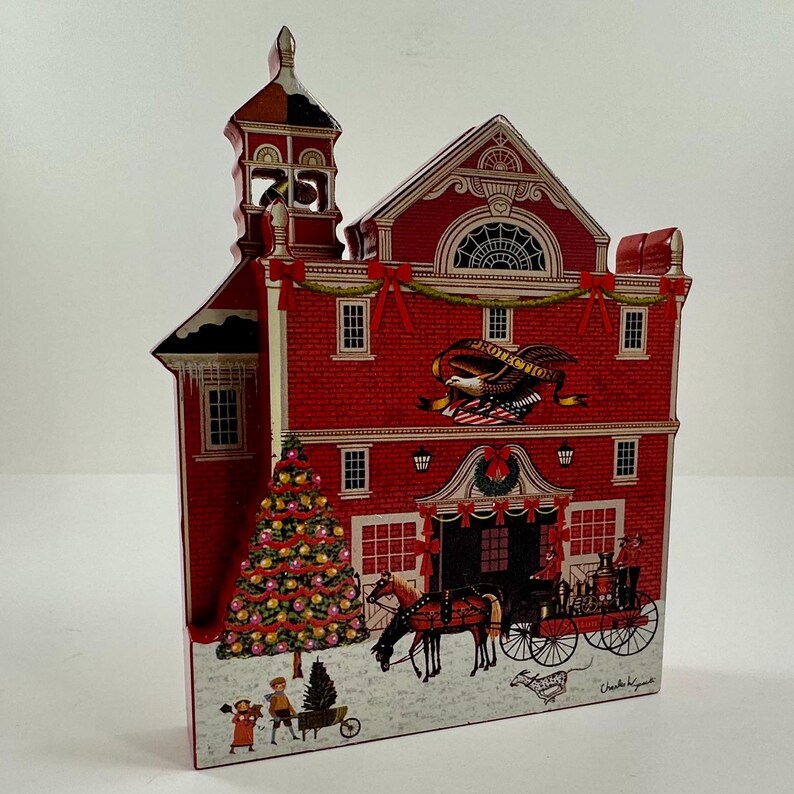 May include: A red wooden Christmas ornament depicting a firehouse with a decorated Christmas tree, a horse-drawn carriage, and two children in front. The firehouse has a bell tower and a sign that reads "Protection".