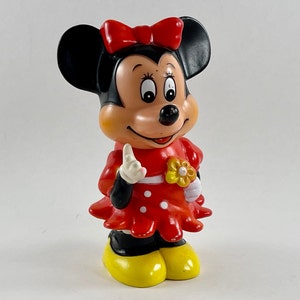 May include: A red and black plastic figurine of Mickey Mouse's girlfriend Minnie Mouse. She is wearing a red dress with white polka dots and a yellow flower. She has a red bow in her hair and is smiling.
