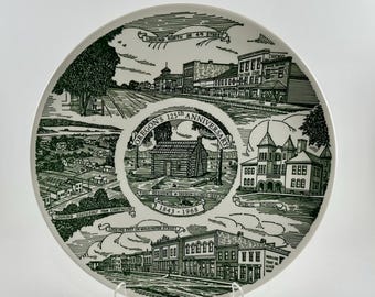 May include: A white ceramic plate with a green illustration commemorating Oregon's 125th anniversary. The plate features historical buildings, a log cabin, and text that reads "Looking Southeast From Courthouse" and "Public School."