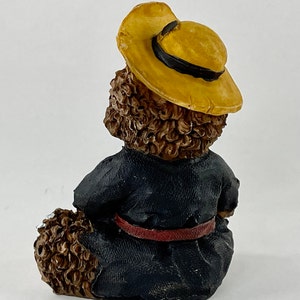 May include: A brown teddy bear figurine wearing a yellow hat and a black robe with a red belt.
