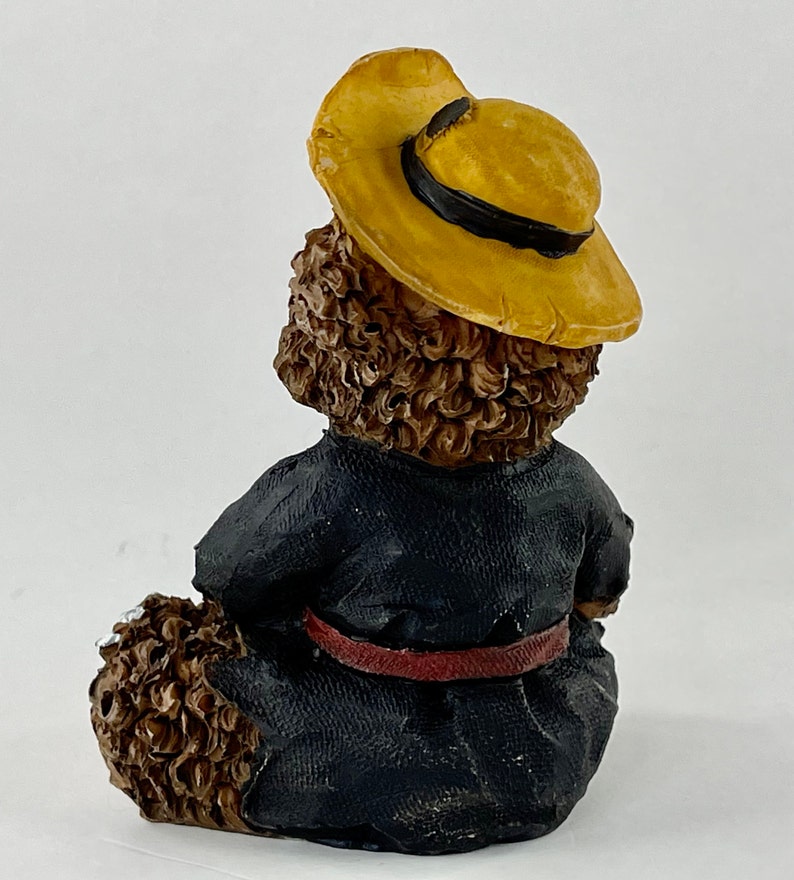 May include: A brown teddy bear figurine wearing a yellow hat and a black robe with a red belt.