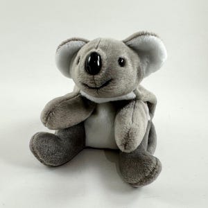 May include: A plush koala bear toy in shades of gray and white. The koala has a black nose, small black eyes, and a smiling mouth. The toy is sitting upright with its arms and legs extended.