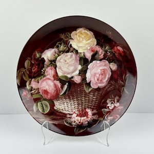 May include: Decorative plate featuring a floral arrangement of pink, red, and yellow roses in a woven basket. The plate has a dark red border and is displayed on a clear stand. A classic decorative piece.