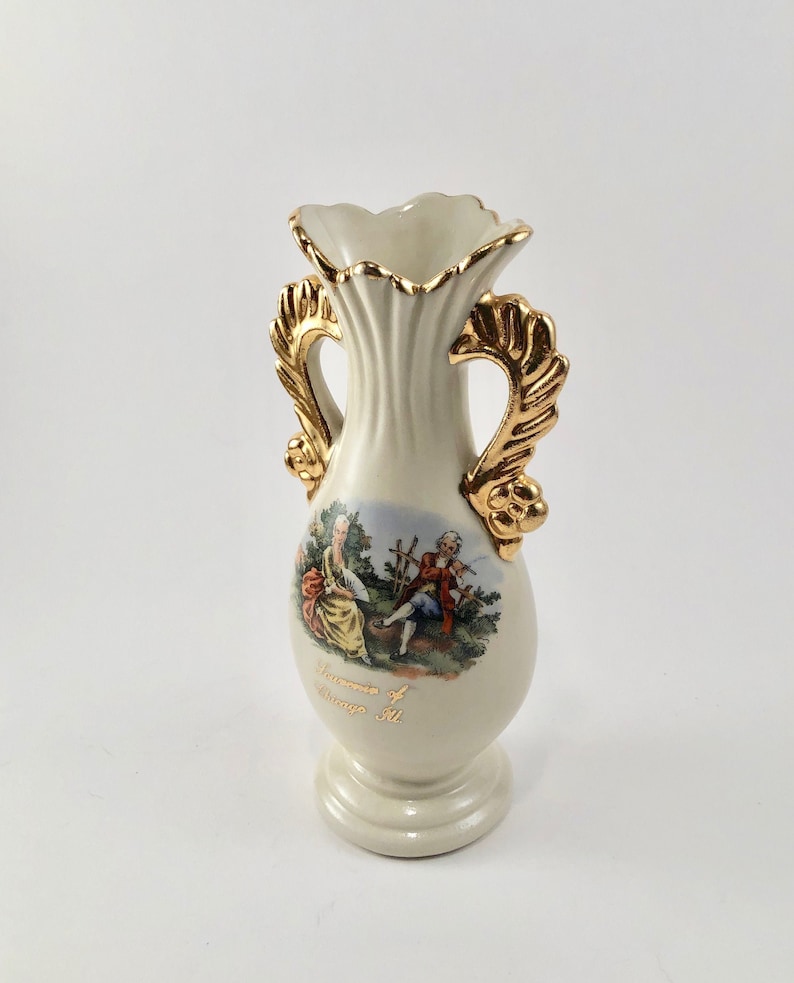 May include: A white ceramic vase with gold accents and a floral design. The vase has two handles and a base. The vase is decorated with a scene of two people in a garden. The text "Souvenirs of Chicago Ill." is printed on the vase.