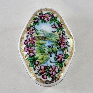 May include: A white porcelain trinket box with a gold trim and a floral design featuring pink roses and blue birds. The box has a landscape scene with a river and a bridge.