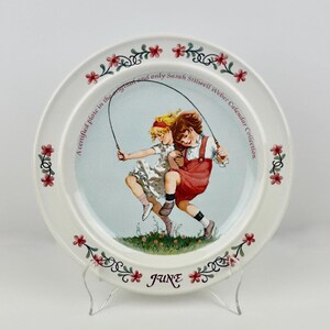 May include: A white decorative plate featuring a vintage illustration of two girls jumping rope. The plate has a floral border with the word "JUNE" and text that reads "A certified plate in the original and only Sarah Stilwell Weber Calendar Collection."