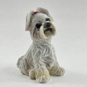 May include: White ceramic figurine of a Maltese dog sitting with its head tilted up. The dog has a red bow on its head.