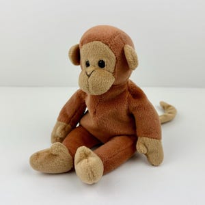 May include: A plush monkey toy in shades of brown and tan. The stuffed animal has a round head with small ears, a tan face, and a long tail. The monkey is sitting with its legs extended.