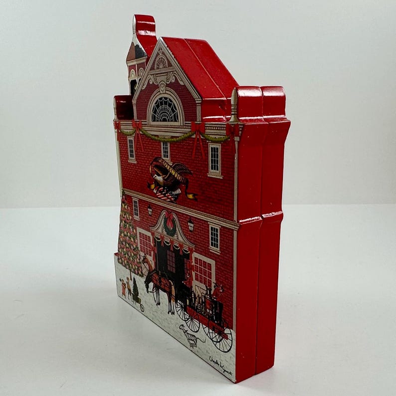 May include: Red tin box shaped like a building with a Christmas tree and a horse-drawn carriage in front. The building has a bell tower and a sign that says "Charles L. Young".