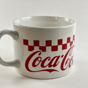 Op de afbeelding: Witte keramische mok met een grijze handgreep, met een rood-wit geblokt patroon en het Coca-Cola-logo in rood schrift. De mok is ontworpen voor dranken en heeft een klassieke, retro esthetiek.