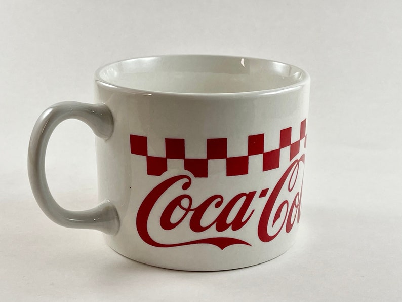 Op de afbeelding: Witte keramische mok met een grijze handgreep, met een rood-wit geblokt patroon en het Coca-Cola-logo in rood schrift. De mok is ontworpen voor dranken en heeft een klassieke, retro esthetiek.