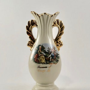 May include: A white ceramic vase with gold accents and handles. The vase features a painted scene of two people in a garden setting. The vase is inscribed with the text "Souvenir of Chicago Ill."