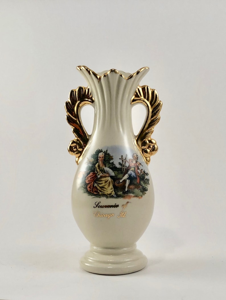 May include: A white ceramic vase with gold accents and handles. The vase features a painted scene of two people in a garden setting. The vase is inscribed with the text "Souvenir of Chicago Ill."