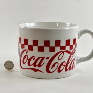 Op de afbeelding: Witte keramische Coca-Cola mok met een rood-wit geblokt patroon en het Coca-Cola logo in rood. De mok heeft een gebogen handvat en staat op een witte achtergrond.