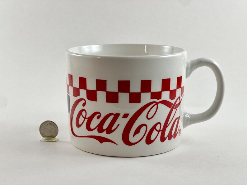 Op de afbeelding: Witte keramische Coca-Cola mok met een rood-wit geblokt patroon en het Coca-Cola logo in rood. De mok heeft een gebogen handvat en staat op een witte achtergrond.
