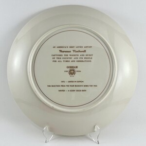 May include: A white ceramic plate with a gold border and inscription. The inscription reads "As America's Best Loved Artist Norman Rockwell Captured the Warmth and Spirit of this Country and its People for All Times and Generations. Gorham Fine USA China 1975 - Limited in Edition This Selection from the Four Season's Series for 1954 Winter - A Lickin' Good Bath"