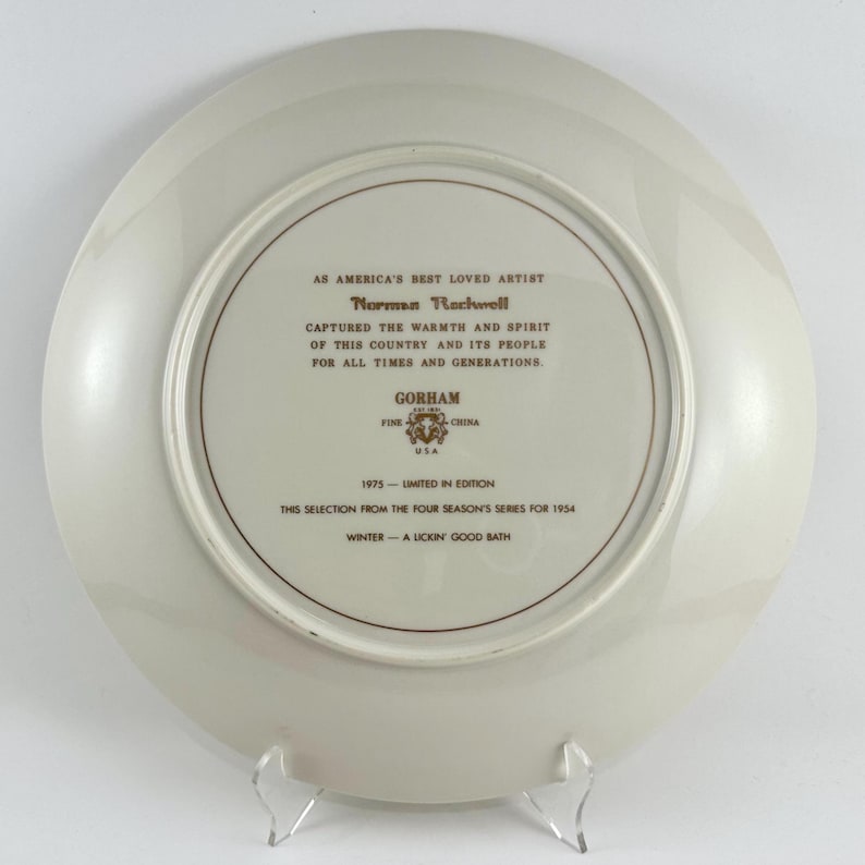 May include: A white ceramic plate with a gold border and inscription. The inscription reads "As America's Best Loved Artist Norman Rockwell Captured the Warmth and Spirit of this Country and its People for All Times and Generations. Gorham Fine USA China 1975 - Limited in Edition This Selection from the Four Season's Series for 1954 Winter - A Lickin' Good Bath"