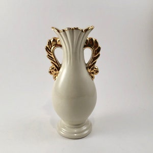 May include: A white ceramic vase with gold leaf accents and two handles. The vase has a fluted design and a rounded body.