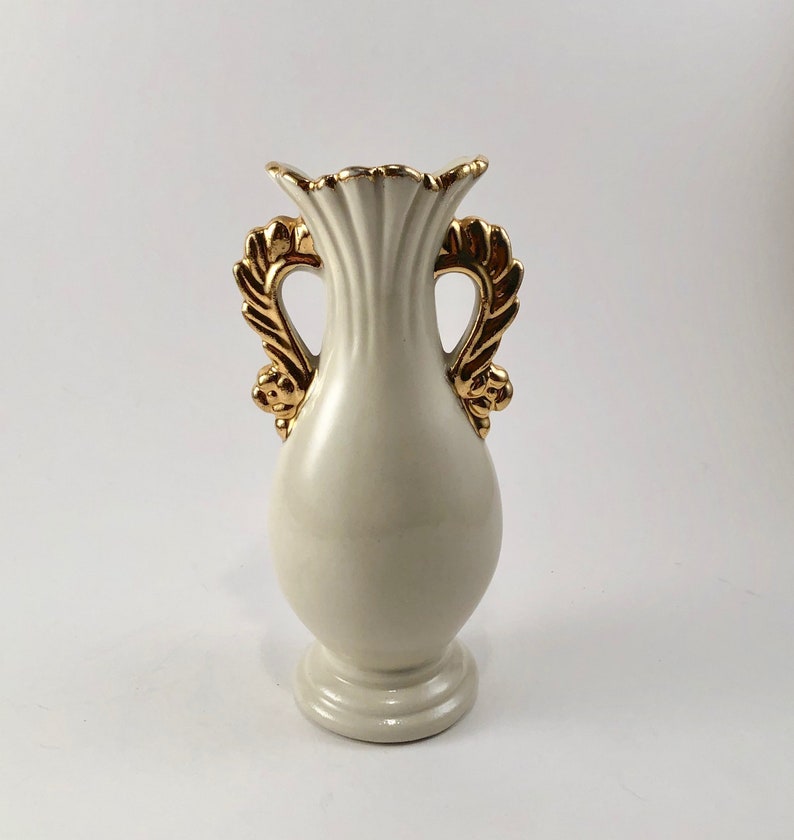 May include: A white ceramic vase with gold leaf accents and two handles. The vase has a fluted design and a rounded body.