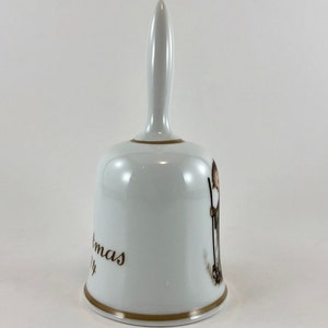 May include: White porcelain Christmas bell with a gold trim and a brown illustration of a person holding a stick. The bell has the words "Christmas" and "Joy" written on it.
