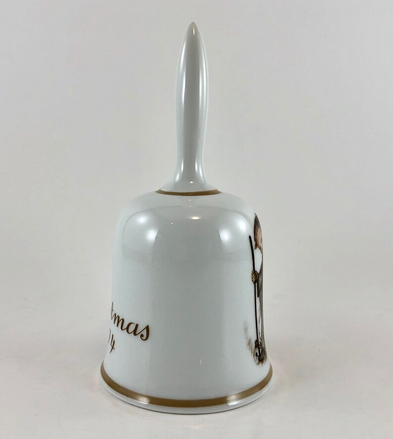 May include: White porcelain Christmas bell with a gold trim and a brown illustration of a person holding a stick. The bell has the words "Christmas" and "Joy" written on it.