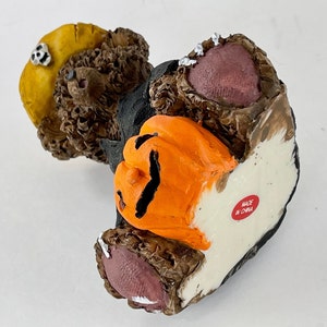 May include: A brown teddy bear wearing a black costume and a yellow hat holds an orange jack-o'-lantern. The bear has a pink nose and pink feet. The bear is made in China.