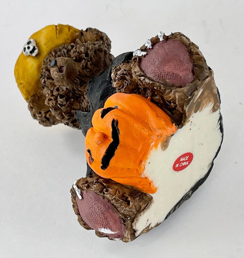 May include: A brown teddy bear wearing a black costume and a yellow hat holds an orange jack-o'-lantern. The bear has a pink nose and pink feet. The bear is made in China.
