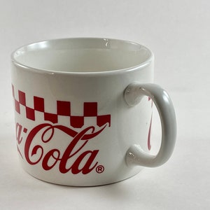 Op de afbeelding: Witte keramische mok met een rood Coca-Cola logo en een rood-wit geblokt patroon. De mok heeft een gebogen handvat. Een klassiek ontwerp voor warme dranken.