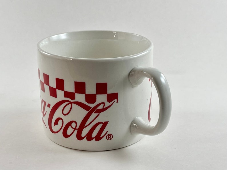 Op de afbeelding: Witte keramische mok met een rood Coca-Cola logo en een rood-wit geblokt patroon. De mok heeft een gebogen handvat. Een klassiek ontwerp voor warme dranken.