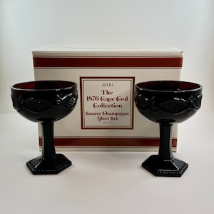 May include: A set of two black glass champagne saucers with a red tint. The saucers are part of the Avon 1876 Cape Cod Collection. The box reads "Saucer Champagne Glass Set (set of 2)"
